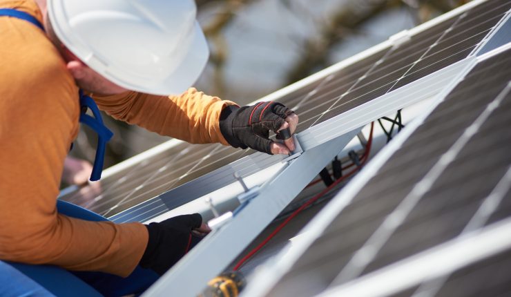 Male workers installing stand-alone solar photovoltaic panel system. Electricians mounting blue solar module on roof of modern house. Alternative energy environmental ecology concept.