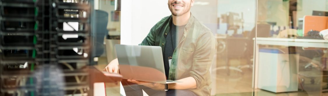 Portrait of happy young male network engineer with laptop in hand working in datacenter.